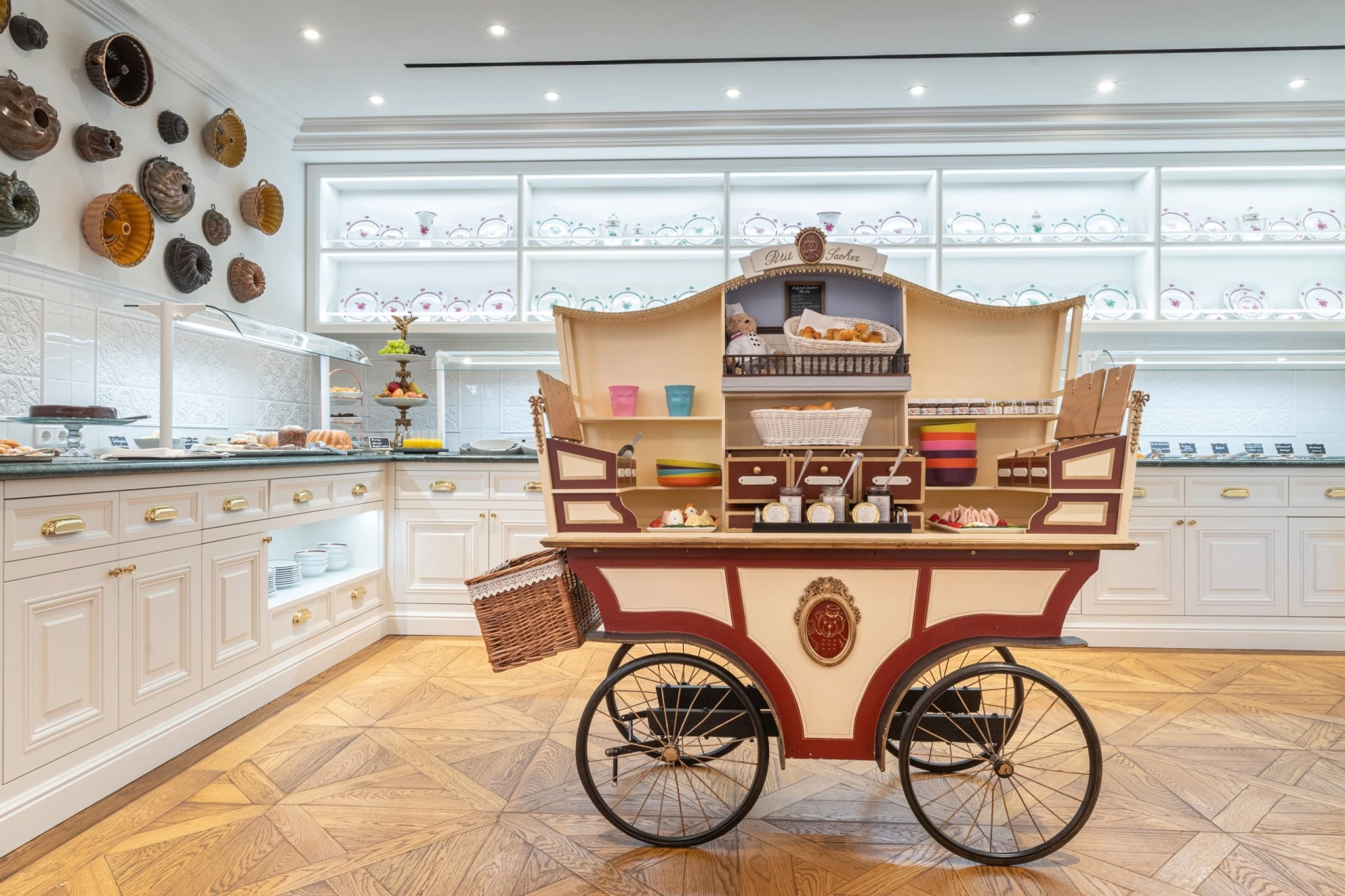 A charming Petit Sacher breakfast cart filled with pastries, jams, and colorful bowls stands in a bright, elegant buffet room at Hotel Sacher, with a teddy bear chef overlooking the delicious offerings.