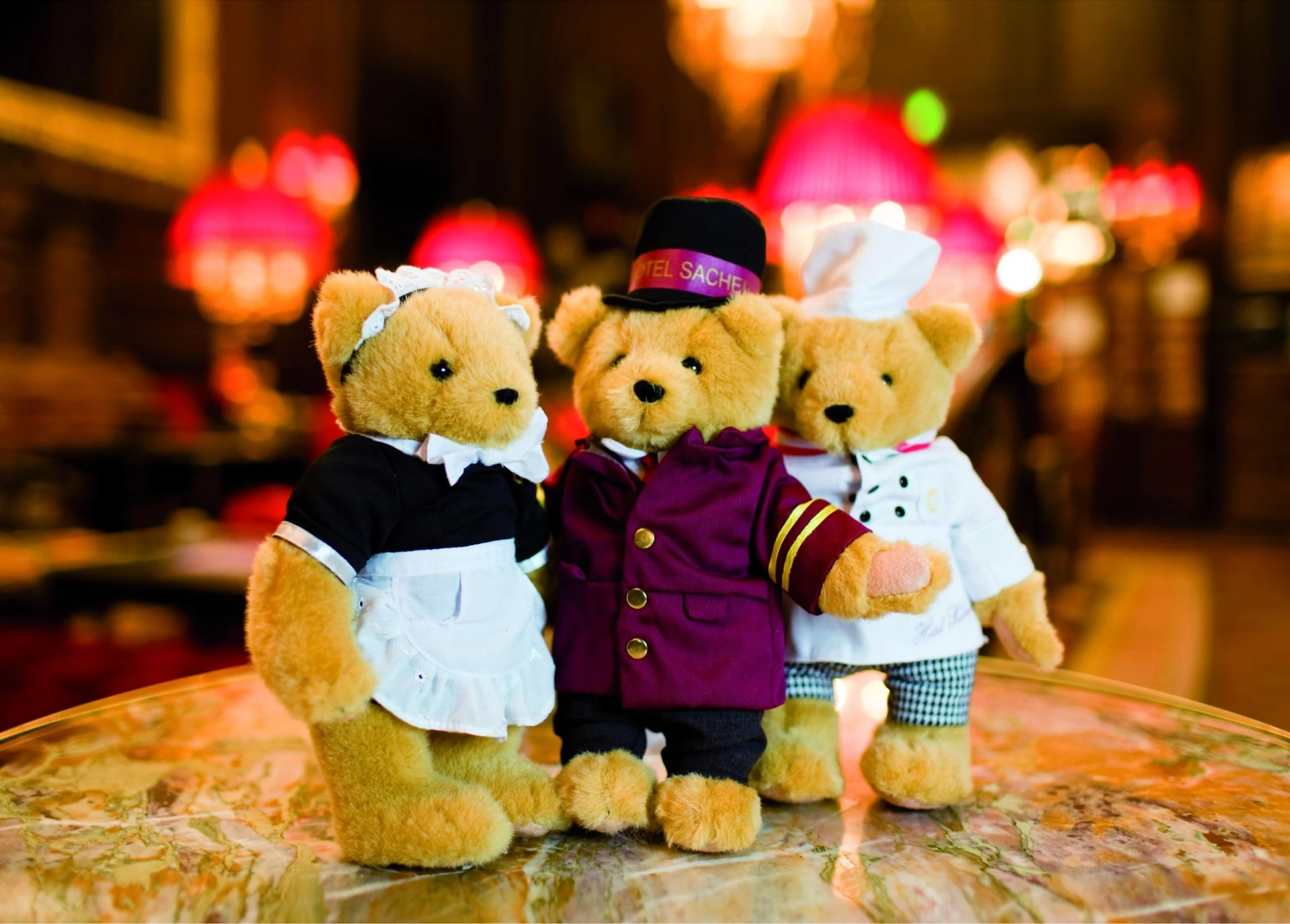 Three teddy bears dressed as Hotel Sacher staff — a maid, a bellboy, and a chef — stand together on a marble table in the opulent lounge of the Hotel Sacher.