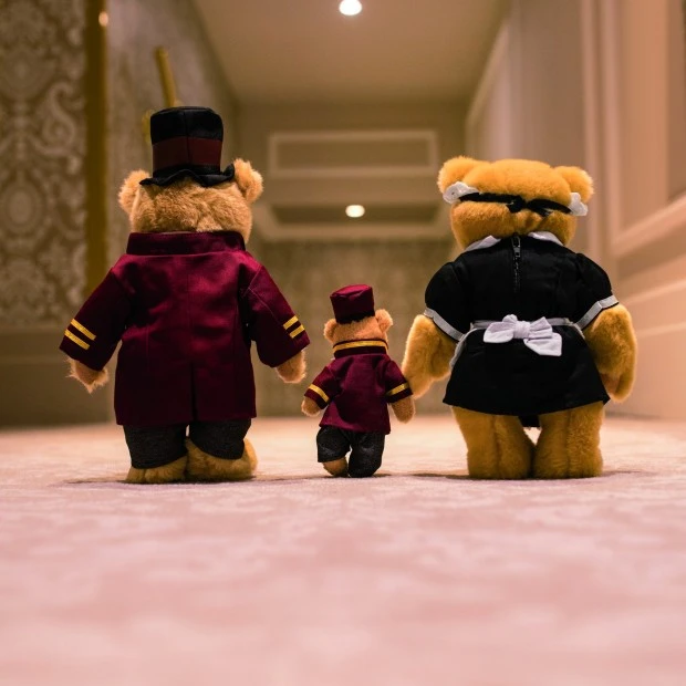 Two teddy bears dressed as Hotel Sacher staff, a bellboy and a maid, walk hand in hand with a smaller teddy bear down a luxurious hallway inside the Hotel Sacher.