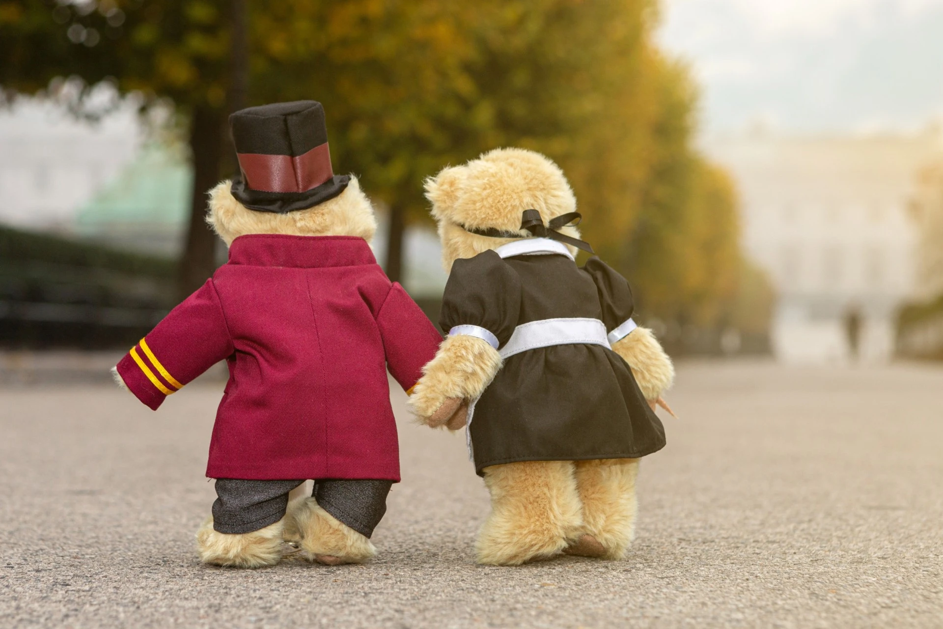 Two teddy bears dressed as a Hotel Sacher bellboy and maid walk hand in hand down a tree-lined avenue, with a grand building softly blurred in the background.