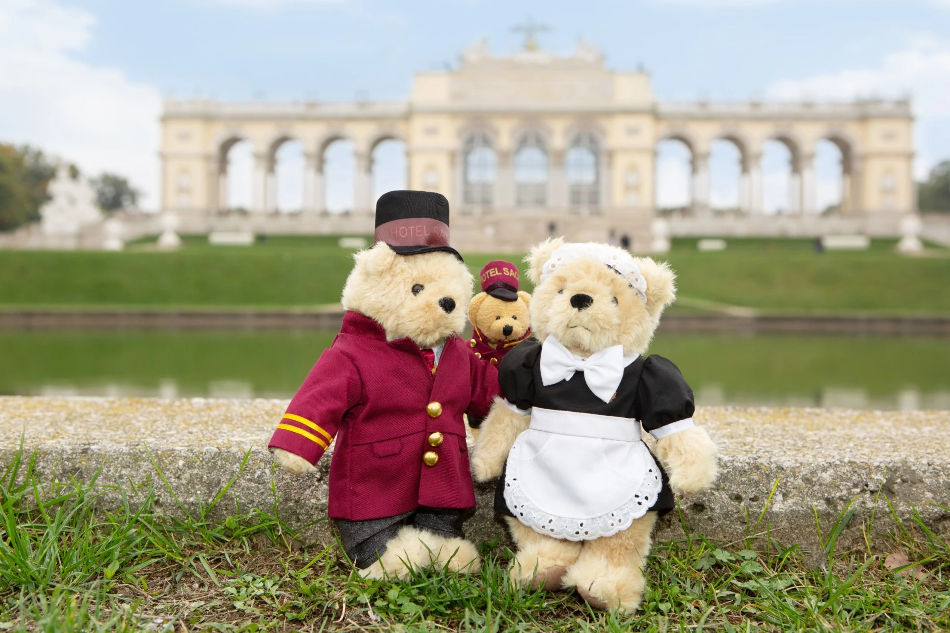 Two teddy bears dressed as Hotel Sacher staff, a bellboy and a maid, along with a smaller teddy bear, pose in front of the Gloriette at Schönbrunn Palace in Vienna.
