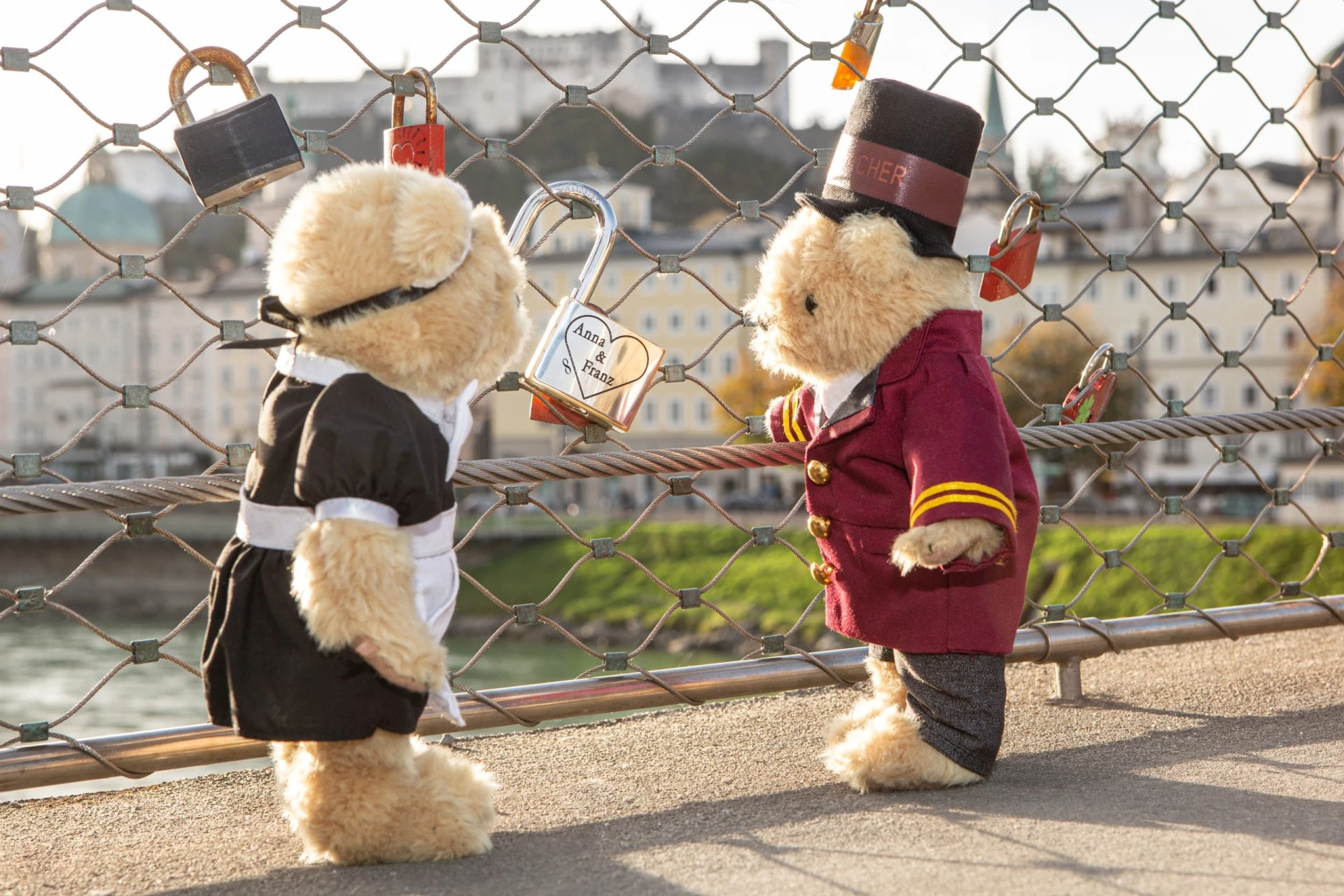 Two teddy bears dressed as Hotel Sacher staff, a maid and a bellboy, stand in front of a love lock fence on a bridge in Salzburg, with a padlock engraved 'Anna & Franz' and the Hohensalzburg Fortress in the background.