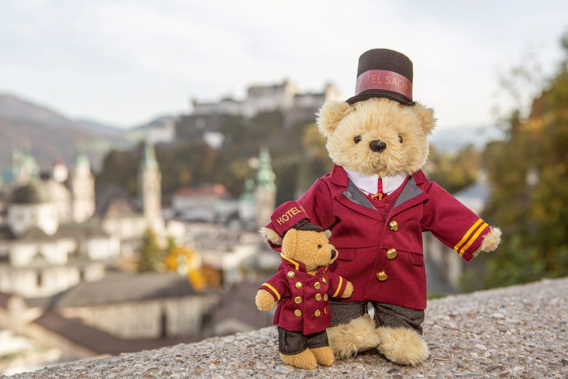 A large and a small teddy bear, both dressed as Hotel Sacher bellboys, stand together overlooking the city of Salzburg with the historic Hohensalzburg Fortress in the background.