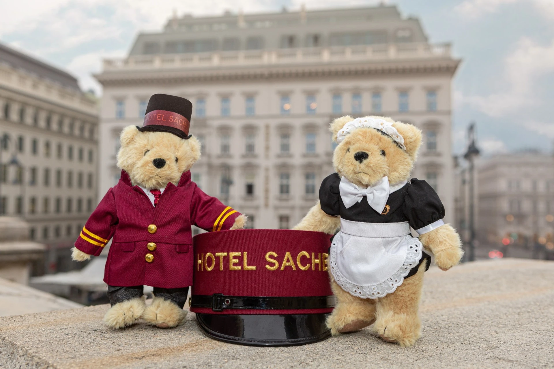Two teddy bears dressed as Hotel Sacher staff, a bellboy and a maid, stand beside a large Hotel Sacher cap with the historic hotel building in the background.