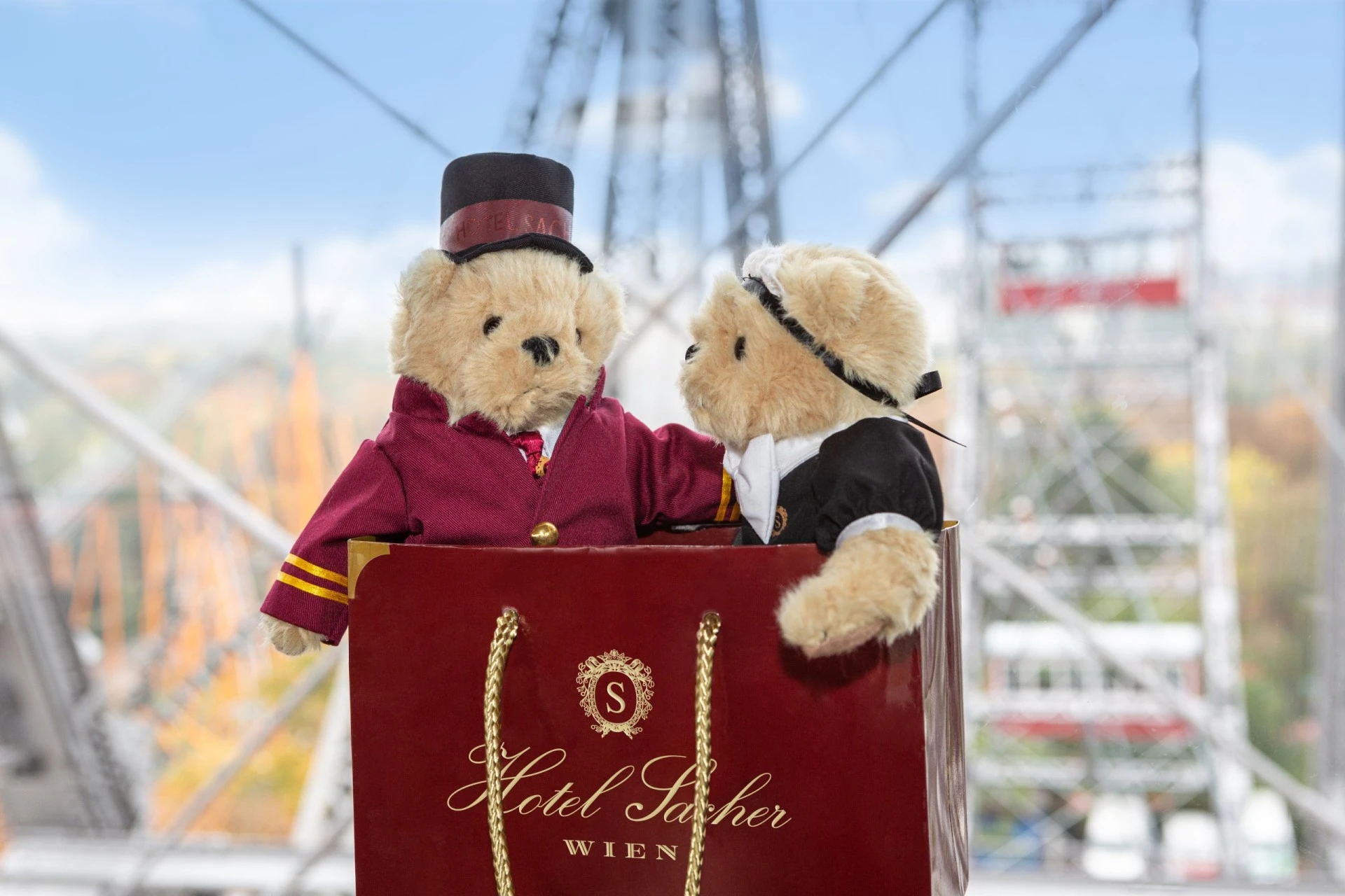 Two plush teddy bears dressed as Hotel Sacher staff sit together inside a red Hotel Sacher Vienna shopping bag, with a Ferris wheel blurred in the background.