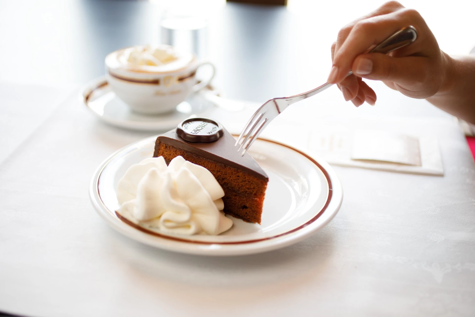 A hand holding a fork reaches for a slice of the Original Sacher-Torte served with a generous portion of whipped cream, alongside a cup of coffee topped with more whipped cream.