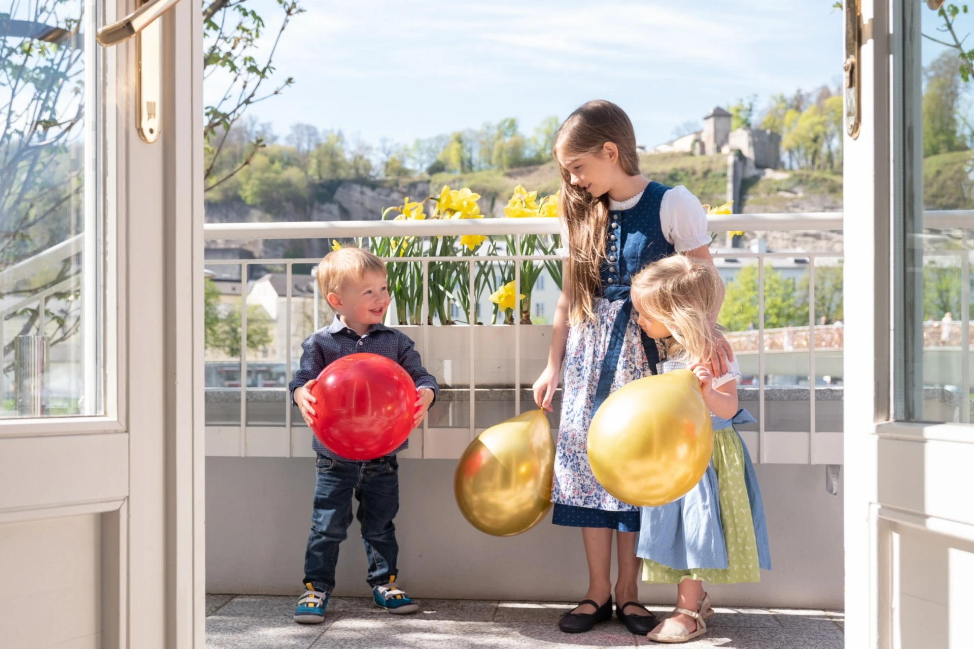 Three children joyfully play with colorful balloons on a sunny balcony, surrounded by blooming daffodils and a charming view of Salzburg in the background.