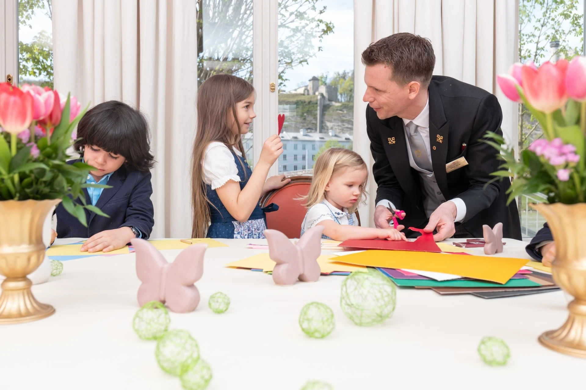 A cheerful crafting session where children are guided through colorful paper creations by a friendly hotel concierge in a festive, flower-filled room.