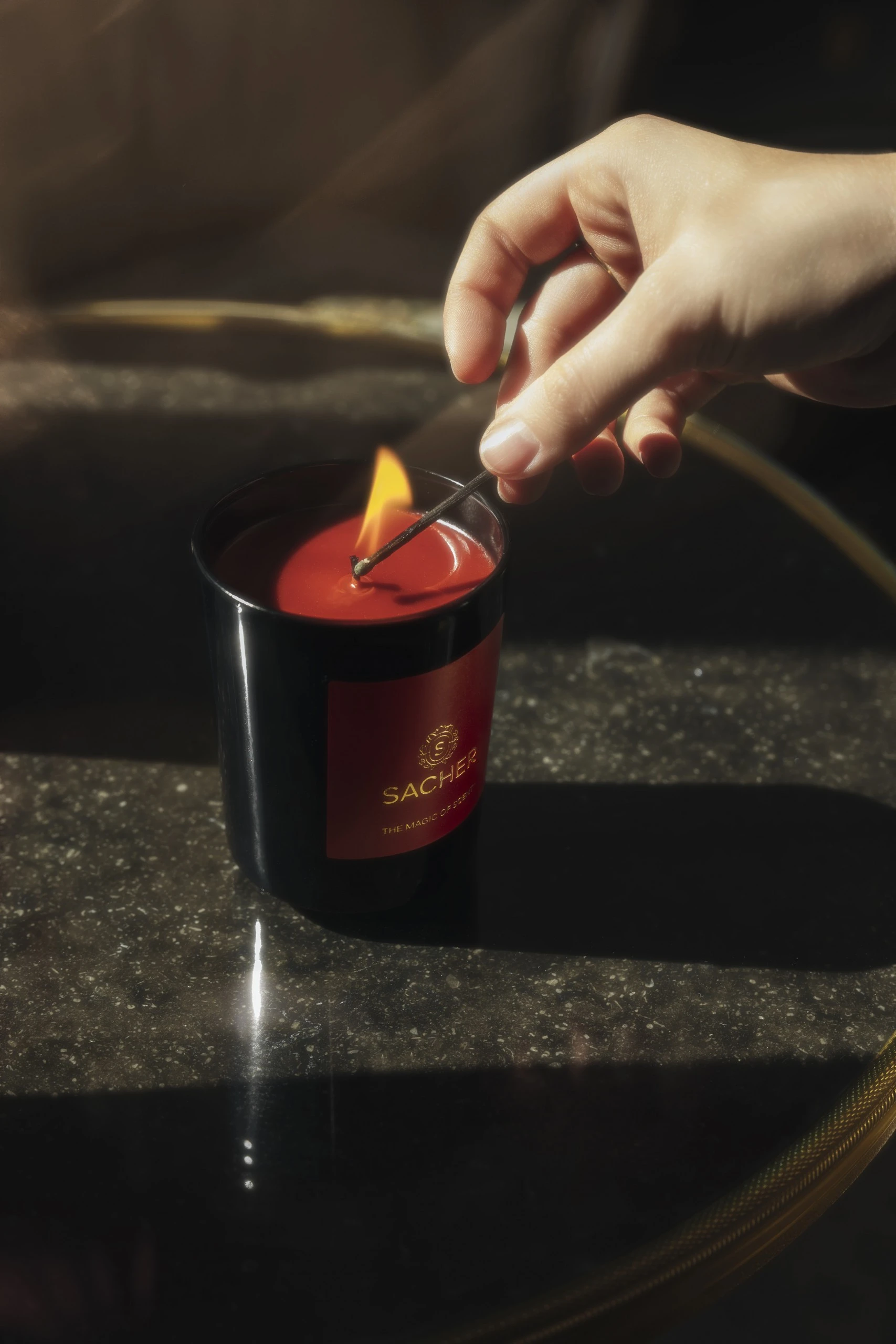 A hand carefully lights a Sacher candle with a match, the soft flame flickering over the deep red wax and casting a warm, golden glow on the dark, textured tabletop below—capturing a quiet moment of indulgent serenity.