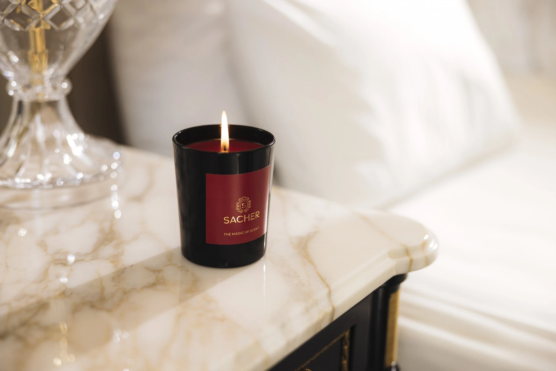 A single Sacher candle with a deep red label glows softly on a polished marble bedside table, casting a warm light beside crisp white linens and a crystal lamp in a luxurious hotel room.