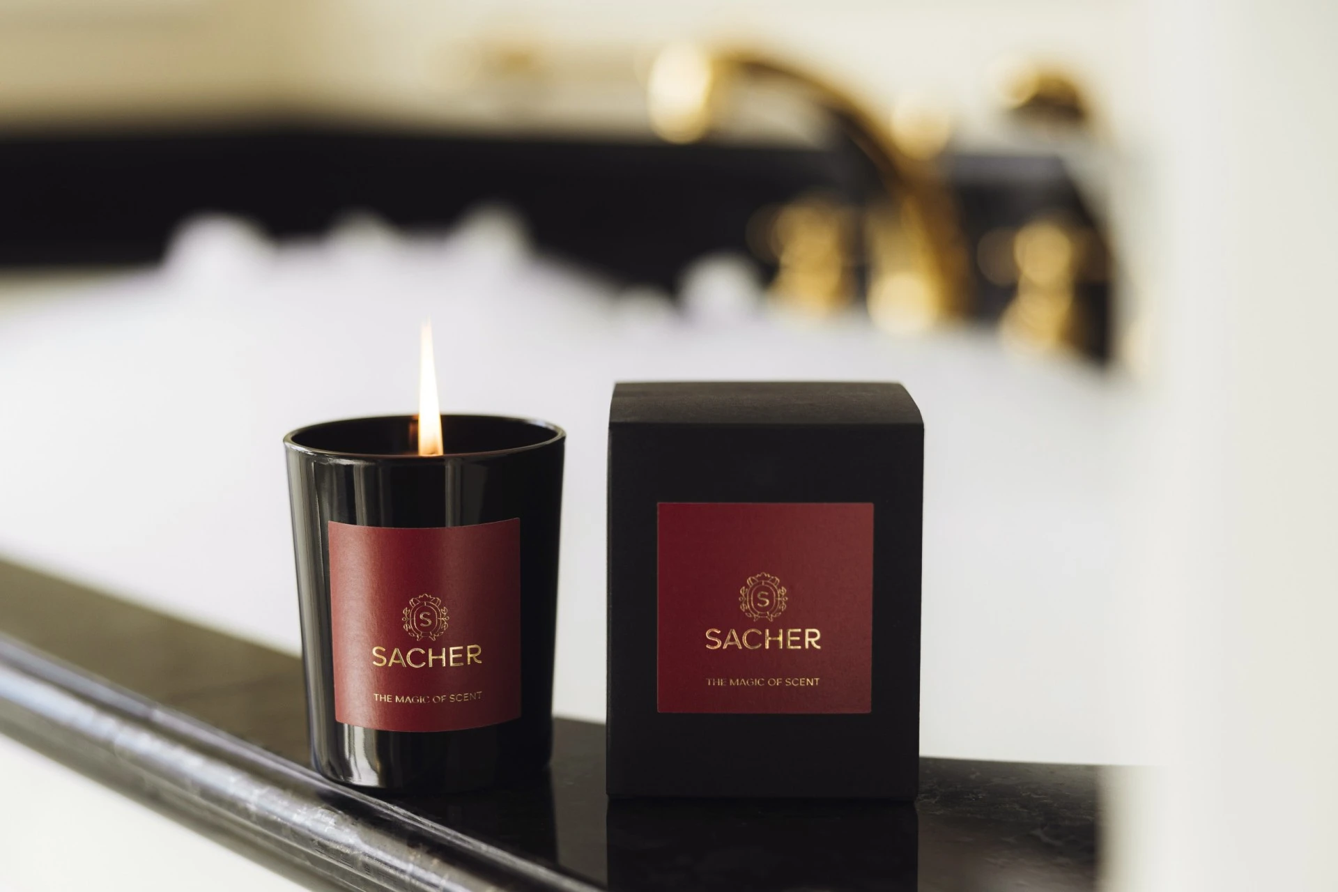A lit Sacher candle with its signature deep red and gold label sits beside its elegant black box on the edge of a luxurious bathtub, with golden fixtures and fluffy white bubbles in the softly blurred background—an invitation to pure relaxation.