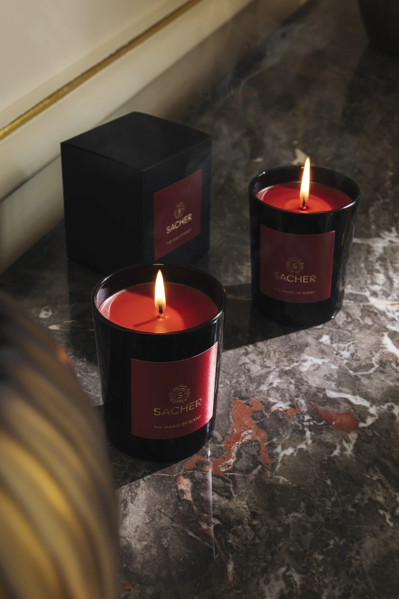 Two elegant black Sacher candles with rich red wax flicker warmly on a polished marble surface, accompanied by a matching box, evoking the luxurious ambiance and signature scent of the Hotel Sacher experience.