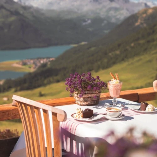 A charming outdoor table set for two features slices of Original Sacher-Torte with whipped cream, cappuccinos, and iced chocolate, all overlooking a breathtaking alpine landscape with a turquoise lake and rolling green mountains.