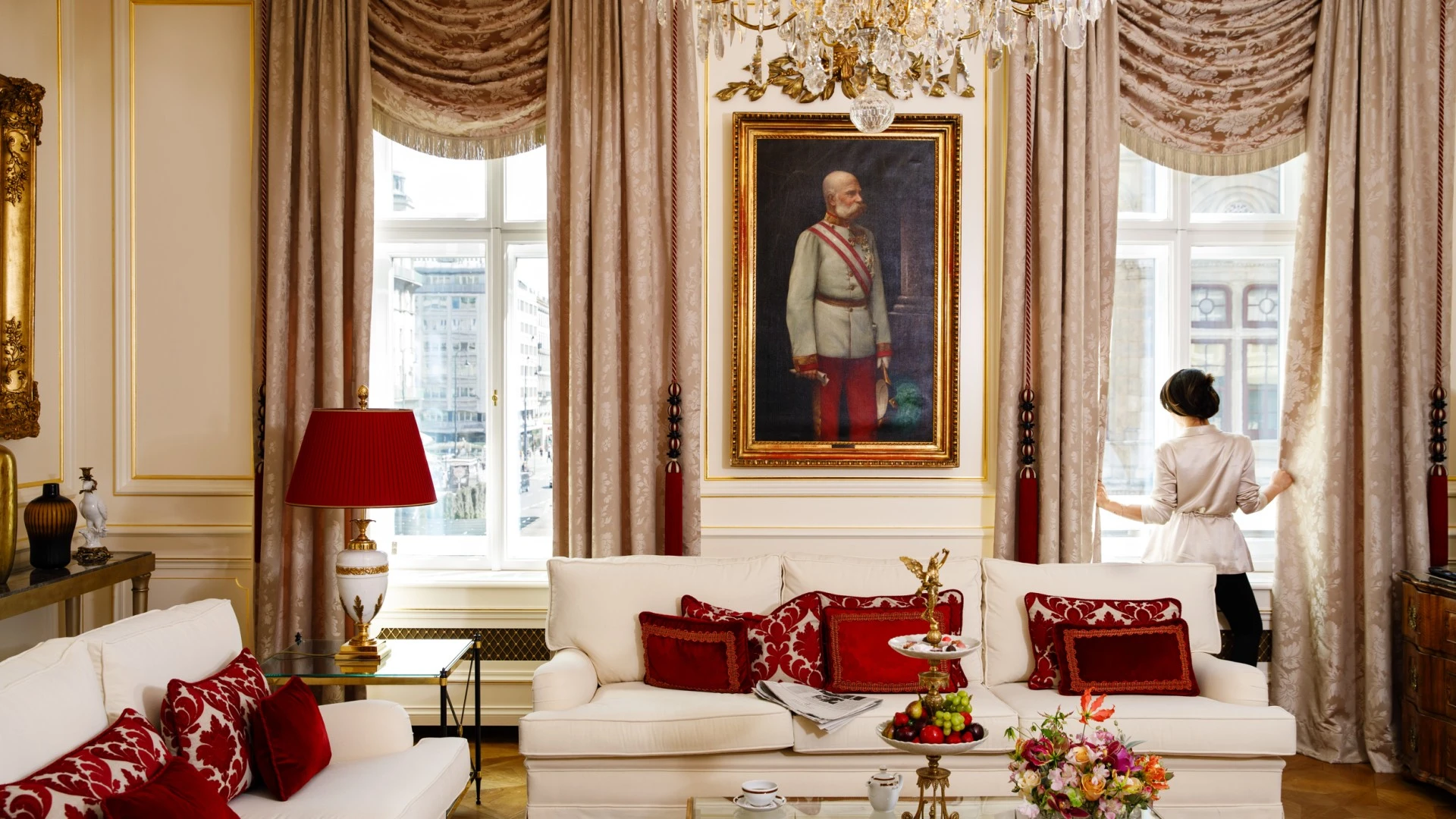 The image depicts an elegant, sunlit living room at Hotel Sacher, richly decorated with cream and red furnishings, a golden chandelier, and a portrait of Emperor Franz Joseph I, as a woman in a satin robe gently opens the curtains to reveal a view of Vienna.