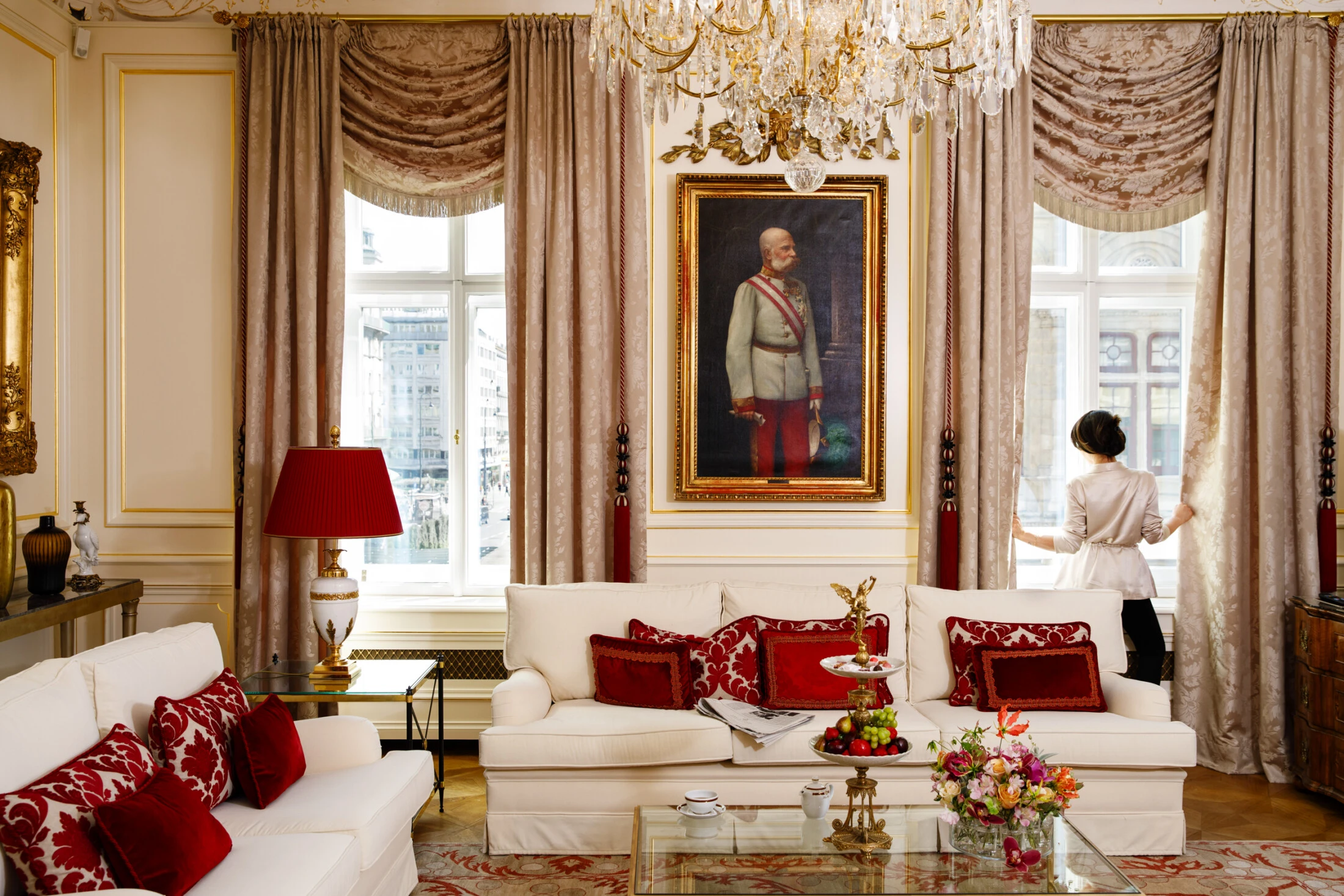 The image depicts an elegant, sunlit living room at Hotel Sacher, richly decorated with cream and red furnishings, a golden chandelier, and a portrait of Emperor Franz Joseph I, as a woman in a satin robe gently opens the curtains to reveal a view of Vienna.