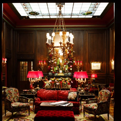 The image shows the opulent lobby of Hotel Sacher, adorned with deep wooden paneling, a grand chandelier, and richly upholstered red and floral furniture, creating a warm and elegant Viennese atmosphere.