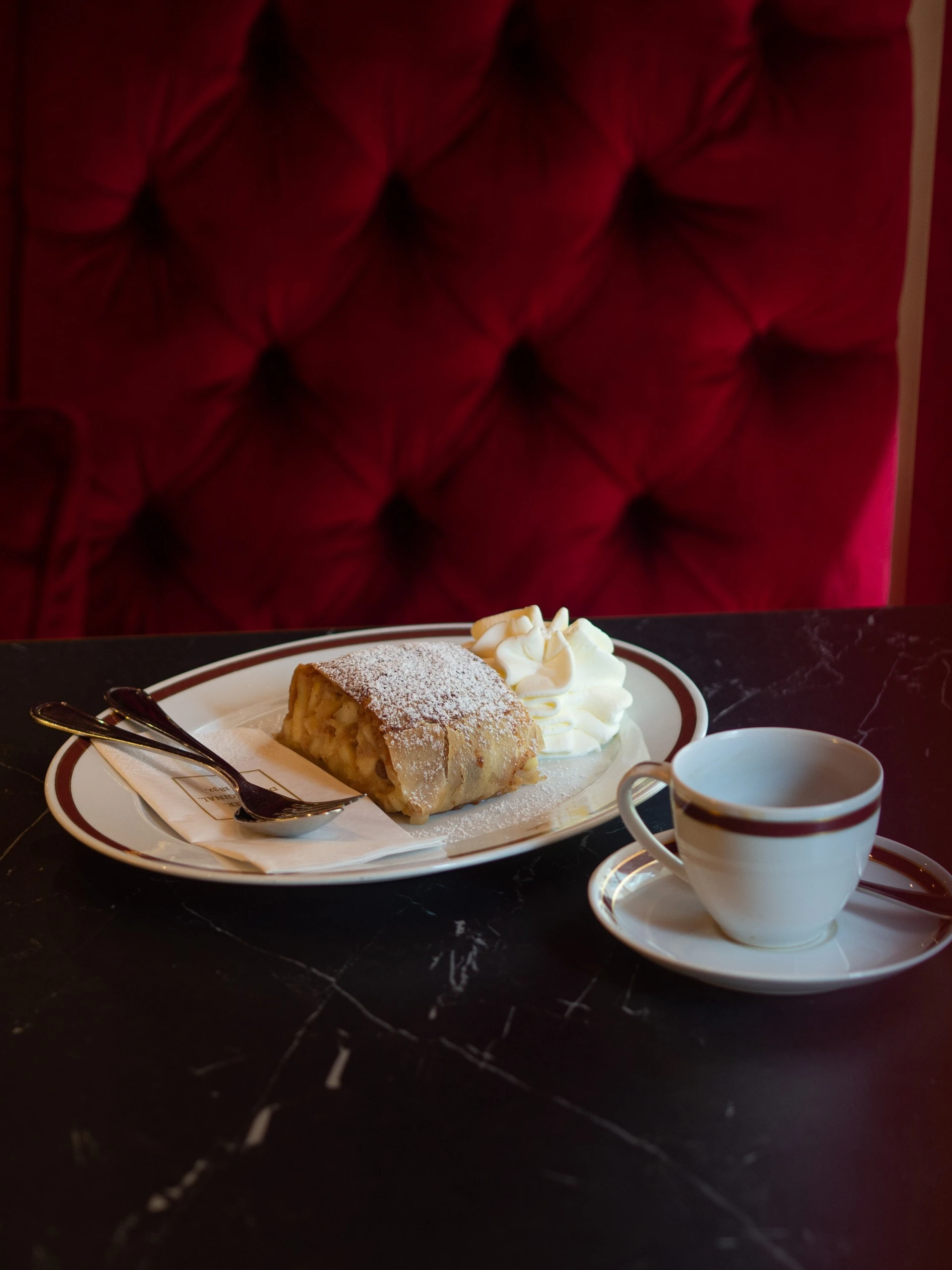 The image shows a slice of traditional apple strudel dusted with powdered sugar, served with a generous dollop of whipped cream and a cup of coffee on elegant Sacher-branded china, set against a plush red velvet backdrop.