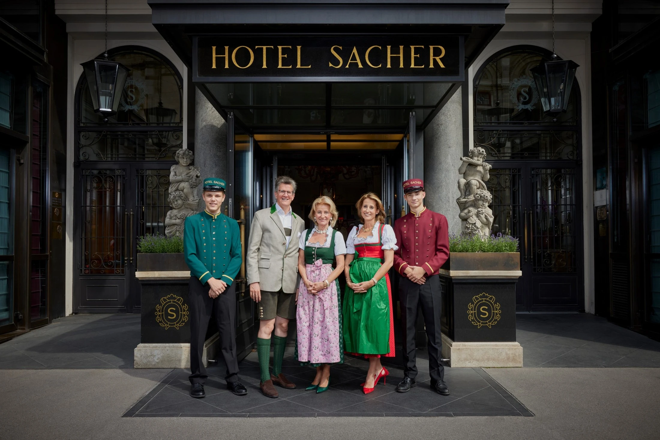In front of the grand entrance to Hotel Sacher, Georg Gürtler, Elisabeth Gürtler, and Alexandra Winkler stand between two uniformed doormen, all smiling, with the family dressed in traditional Austrian attire.