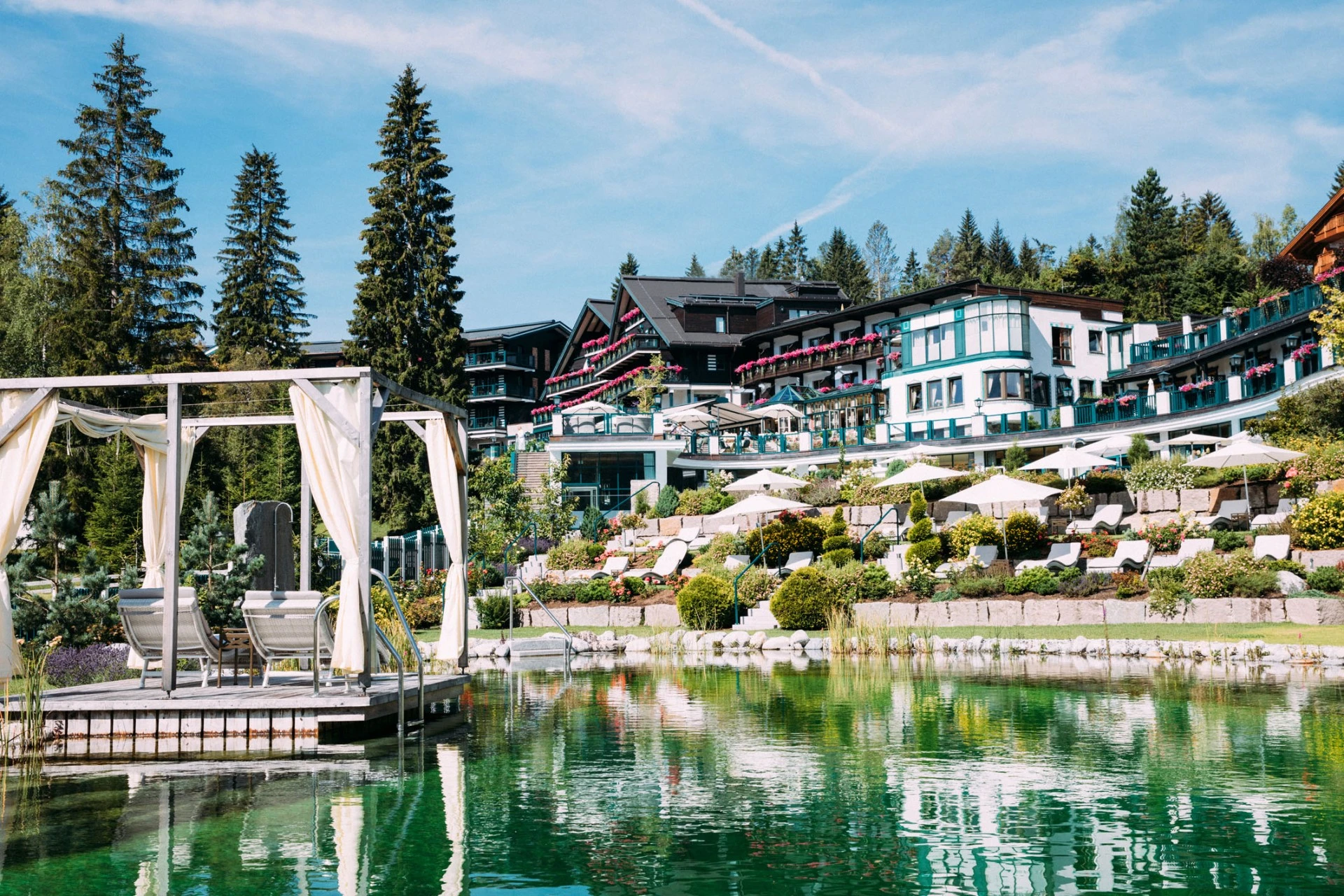 This photo captures the idyllic alpine elegance of Hotel Sacher Seefeld, nestled in the Tyrolean mountains with its tranquil natural pool, lush landscaped gardens, and luxurious terraces framed by tall pine trees and vibrant floral balconies.