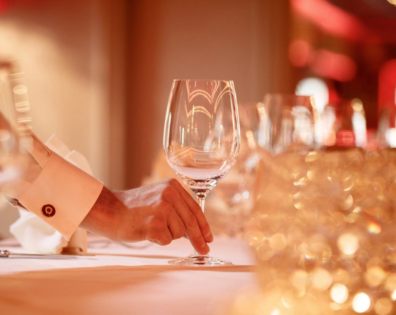 The image shows an elegant hand delicately placing a wine glass on a beautifully set table, bathed in warm ambient lighting that evokes a sense of refined luxury and sophistication.