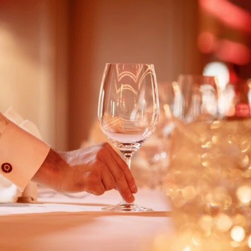 The image shows an elegant hand delicately placing a wine glass on a beautifully set table, bathed in warm ambient lighting that evokes a sense of refined luxury and sophistication.