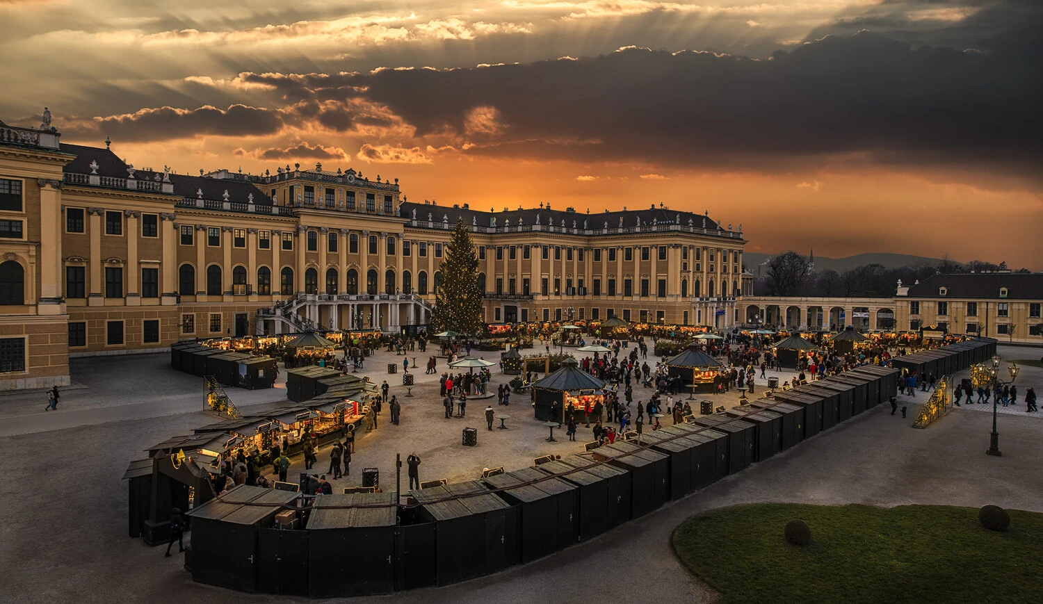 The photo captures a festive Christmas market set against the majestic backdrop of Schönbrunn Palace at sunset, with glowing lights, a towering Christmas tree, and bustling holiday stalls filled with joyful visitors.