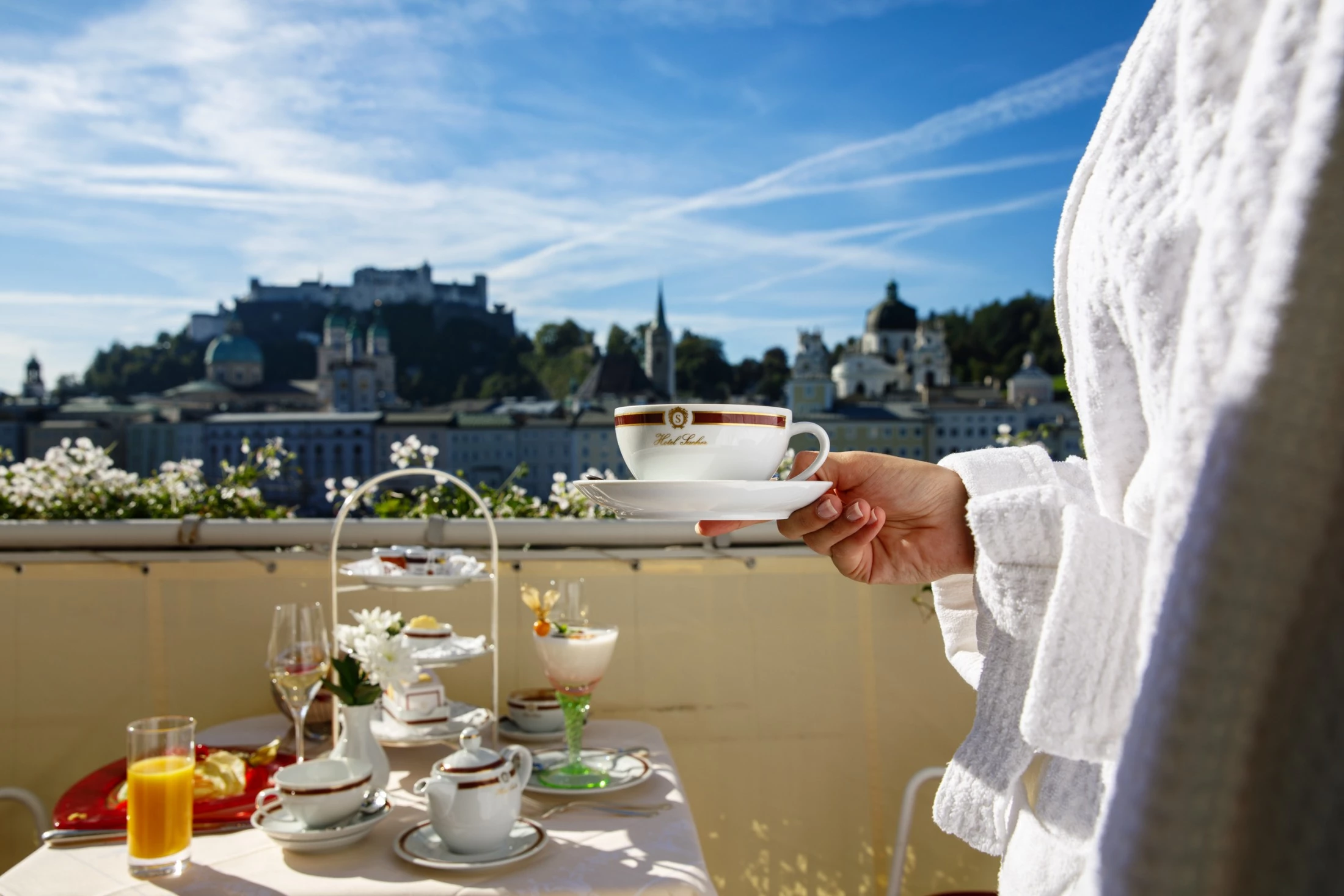 A hand elegantly holds a porcelain coffee cup on a sunlit terrace, overlooking the historic skyline of Salzburg with a beautifully set breakfast table in the foreground.