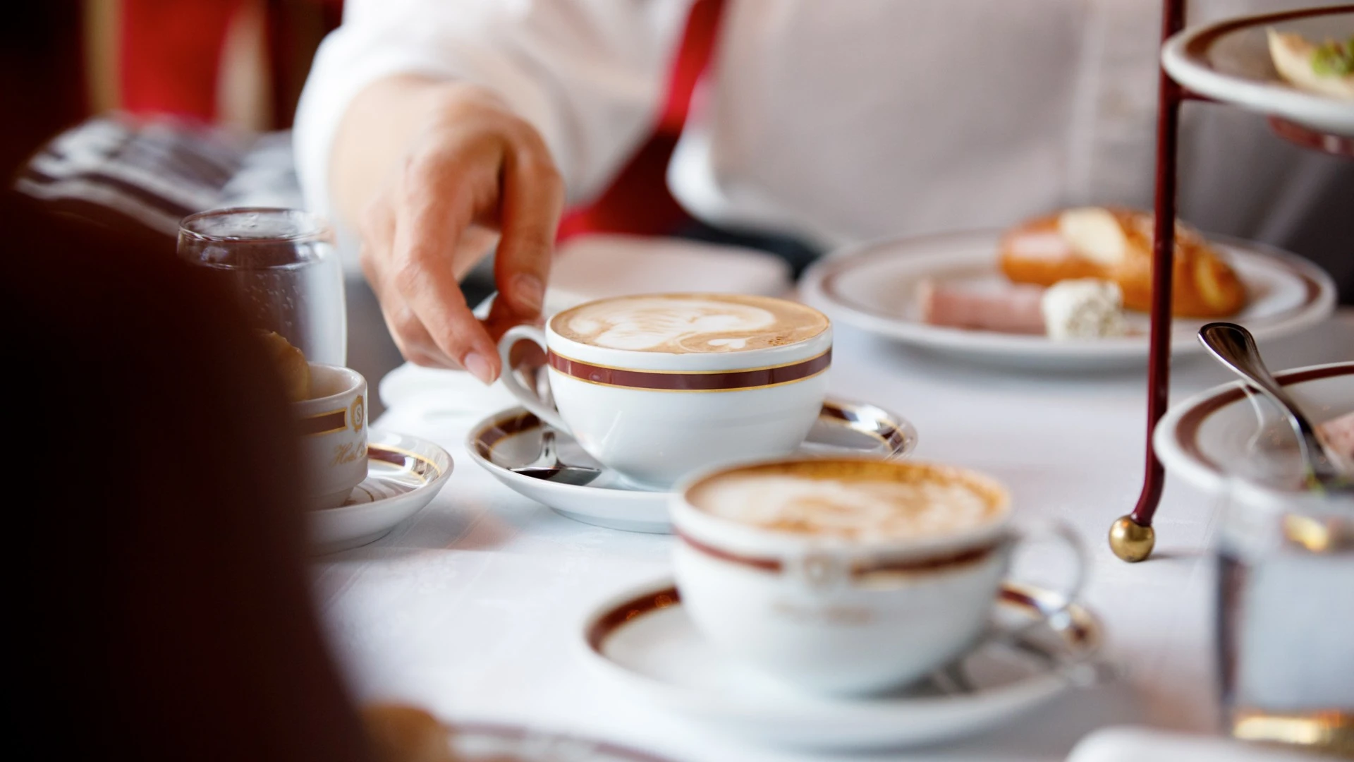 This elegant breakfast scene at Café Sacher exudes classic Viennese charm—delicate porcelain cups trimmed with gold cradle artfully poured cappuccinos, accompanied by a traditional breakfast spread on fine china. The ambiance whispers of quiet luxury, where each moment is savored in style.