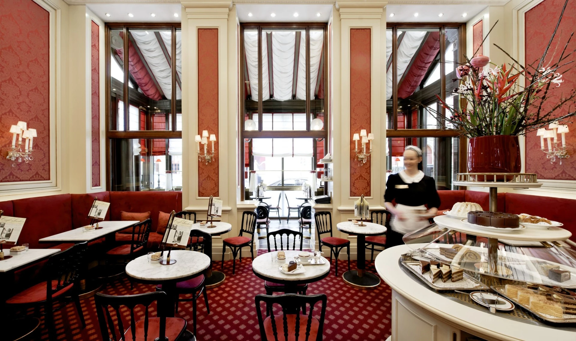 This is the refined interior of a Sacher Café, with tall windows draped in white, red-upholstered seating, delicate wall sconces, and a central pastry display crowned with a grand floral arrangement.
