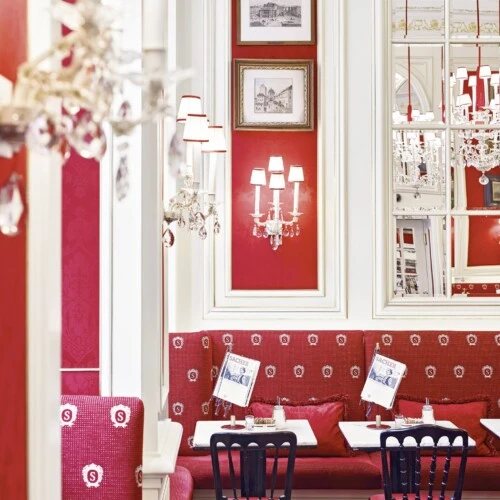 This is a bright and elegant corner of Café Sacher, with signature red seating embroidered with the Sacher emblem, gleaming chandeliers, mirrored walls, and neatly set marble tables ready for guests.