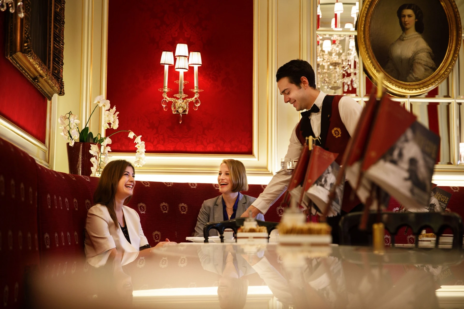 This is a classic Sacher Café scene, where two smiling guests are being warmly served by a traditionally dressed waiter amid rich red décor, golden accents, and an elegant portrait of Empress Elisabeth on the wall.