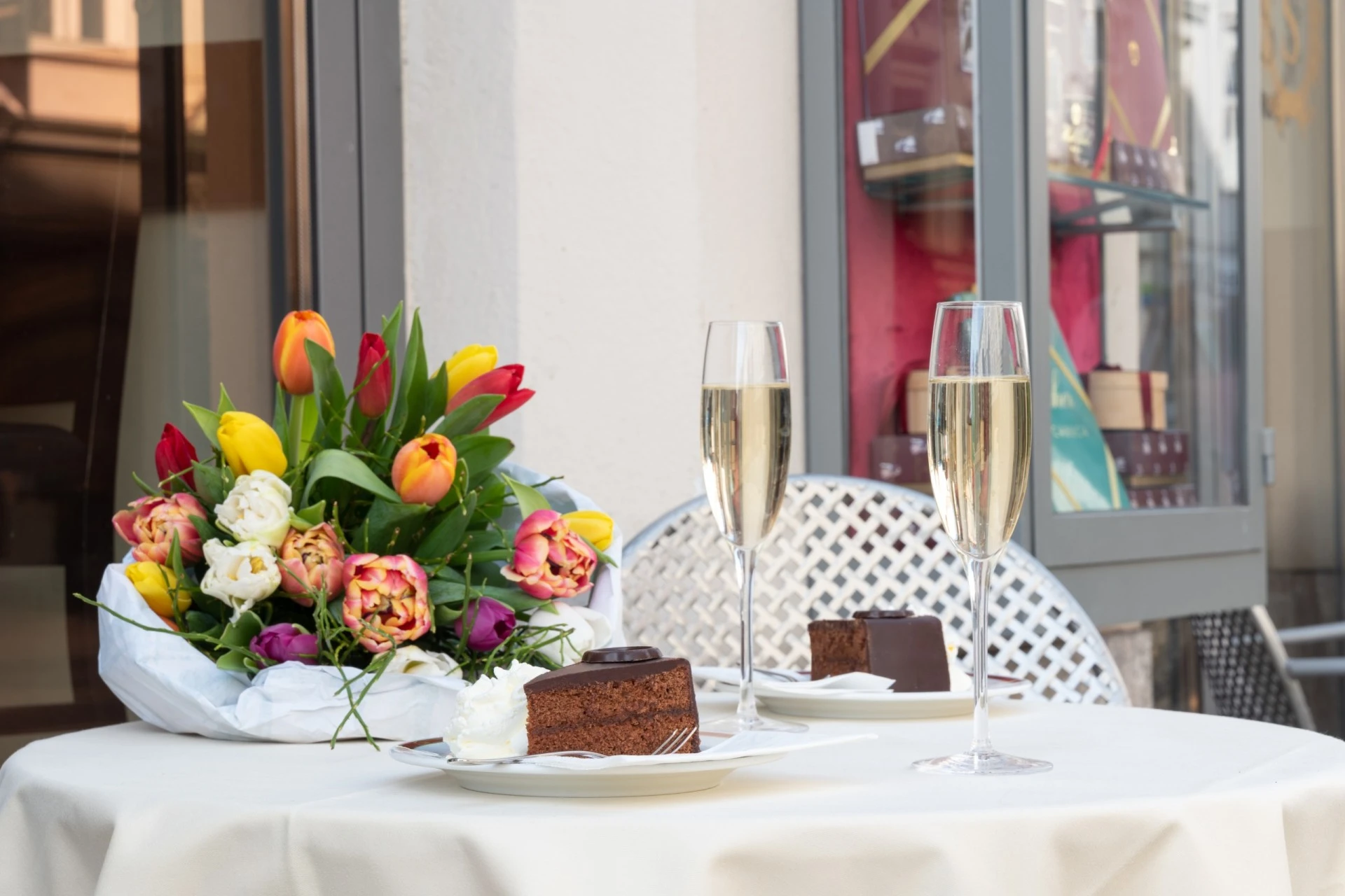 This is a charming outdoor table at Café Sacher Graz, set with two slices of Original Sacher-Torte, glasses of sparkling wine, and a vibrant bouquet of tulips, perfect for a celebratory moment.