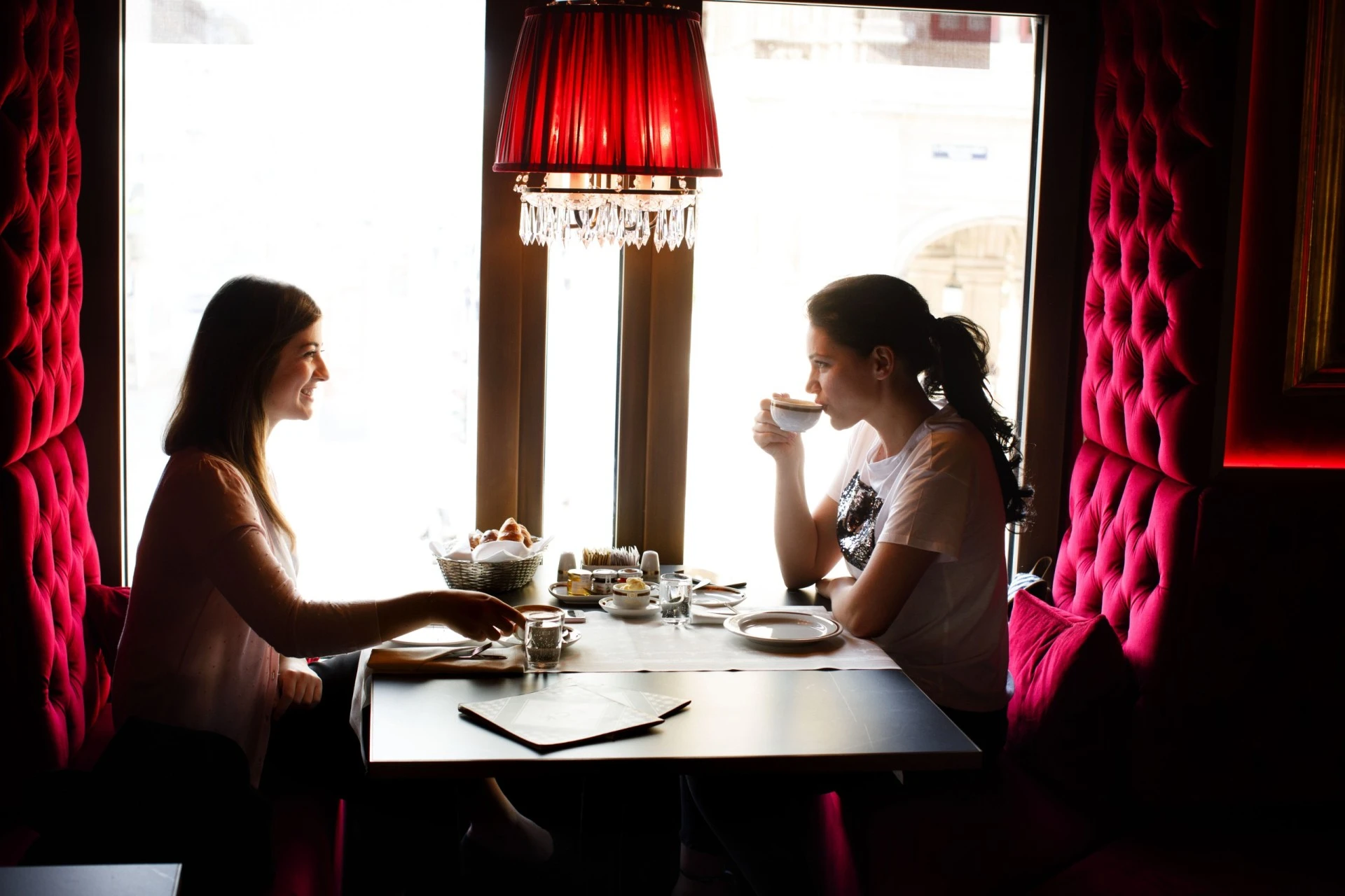This is Restaurant Bel Etage, where two women enjoy a cozy, elegant breakfast by a large window, seated on plush red velvet banquettes beneath a sparkling red chandelier.