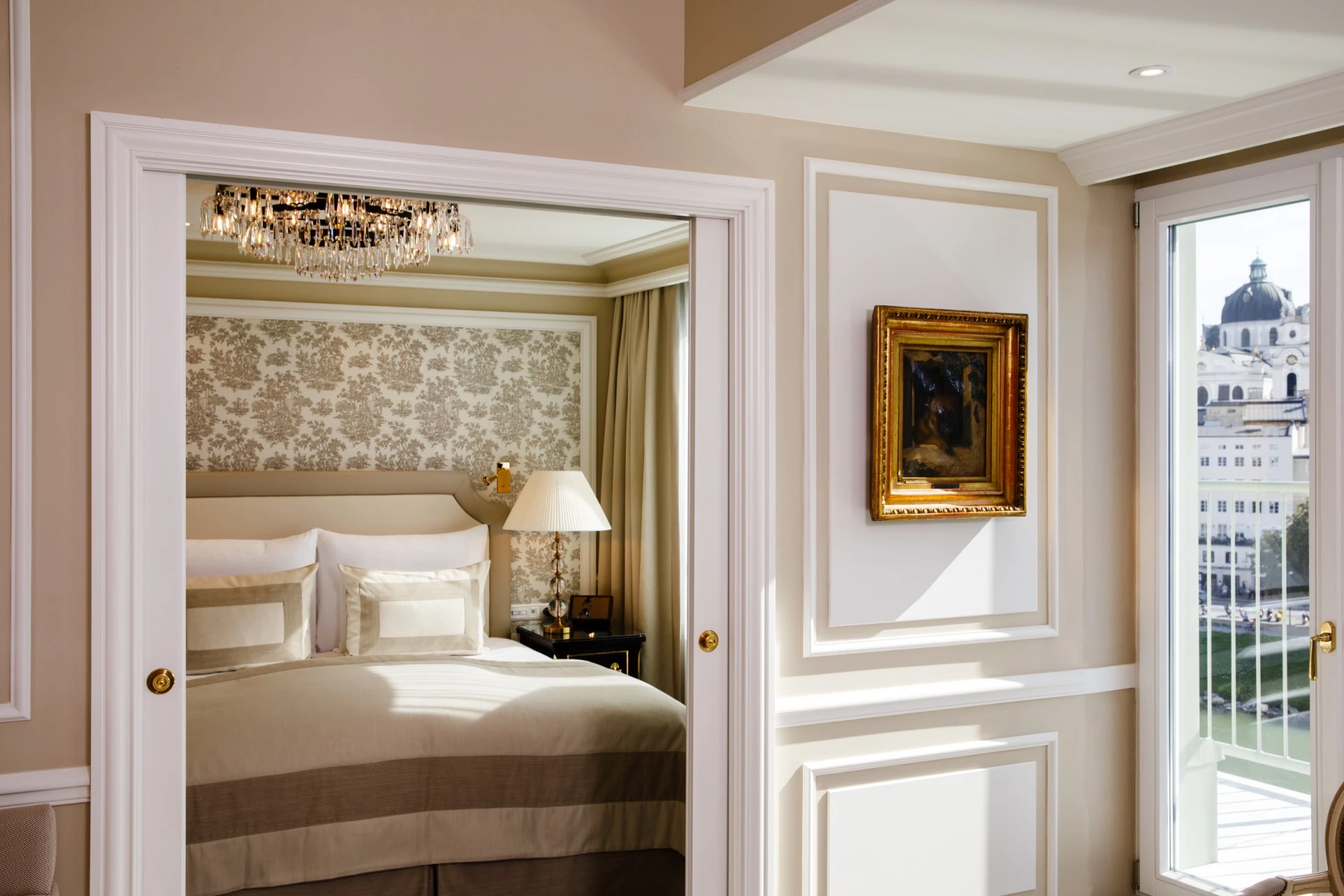 This is a beautifully appointed bedroom at Hotel Sacher Salzburg, featuring elegant cream and gold tones, a sparkling crystal chandelier, and a view of Salzburg’s historic domes through tall glass doors.