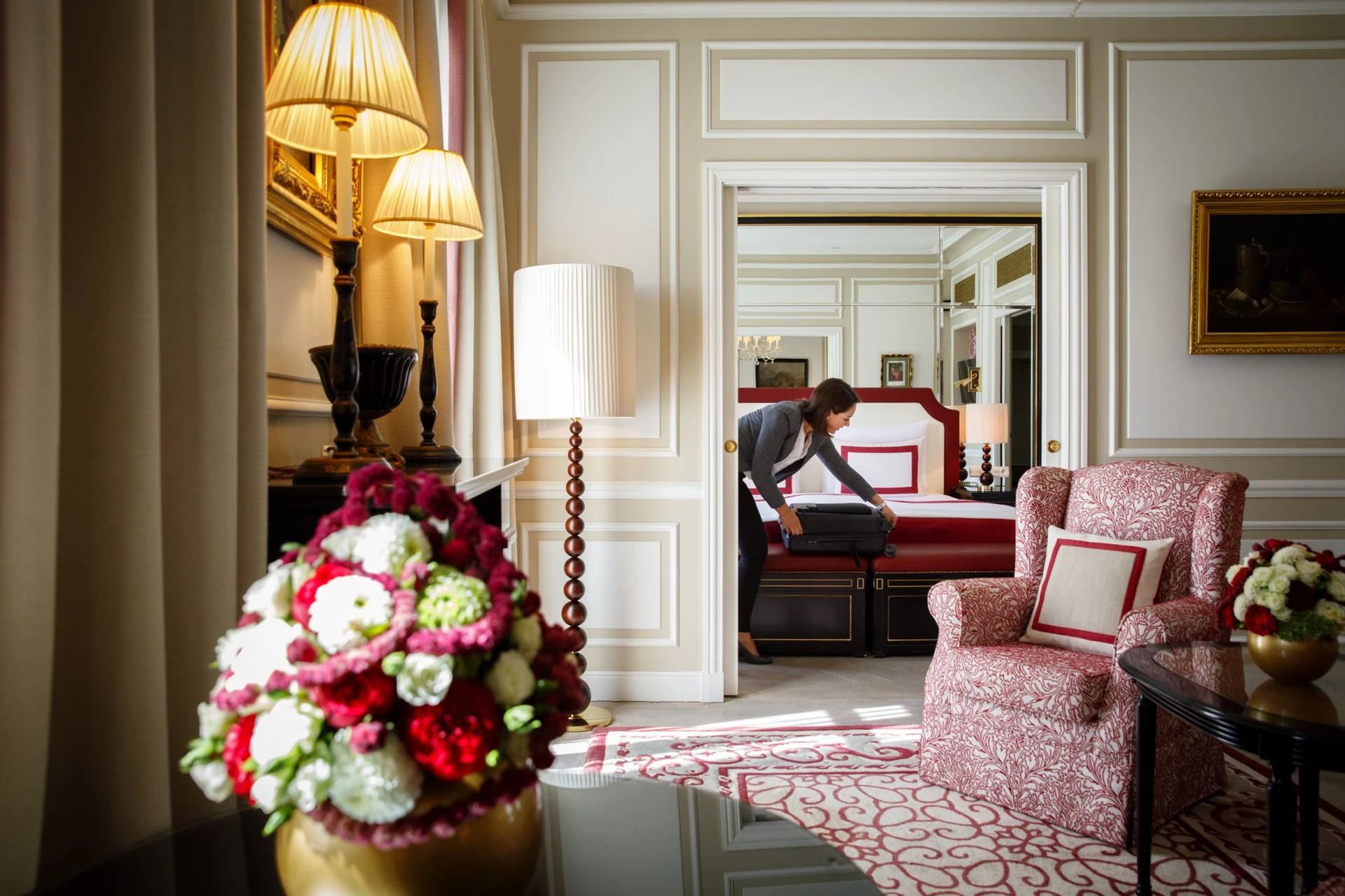 This is a refined and opulent room at Hotel Sacher Salzburg, with elegant classic furnishings, rich red and cream tones, and a guest preparing her stay in the plush bedroom beyond.