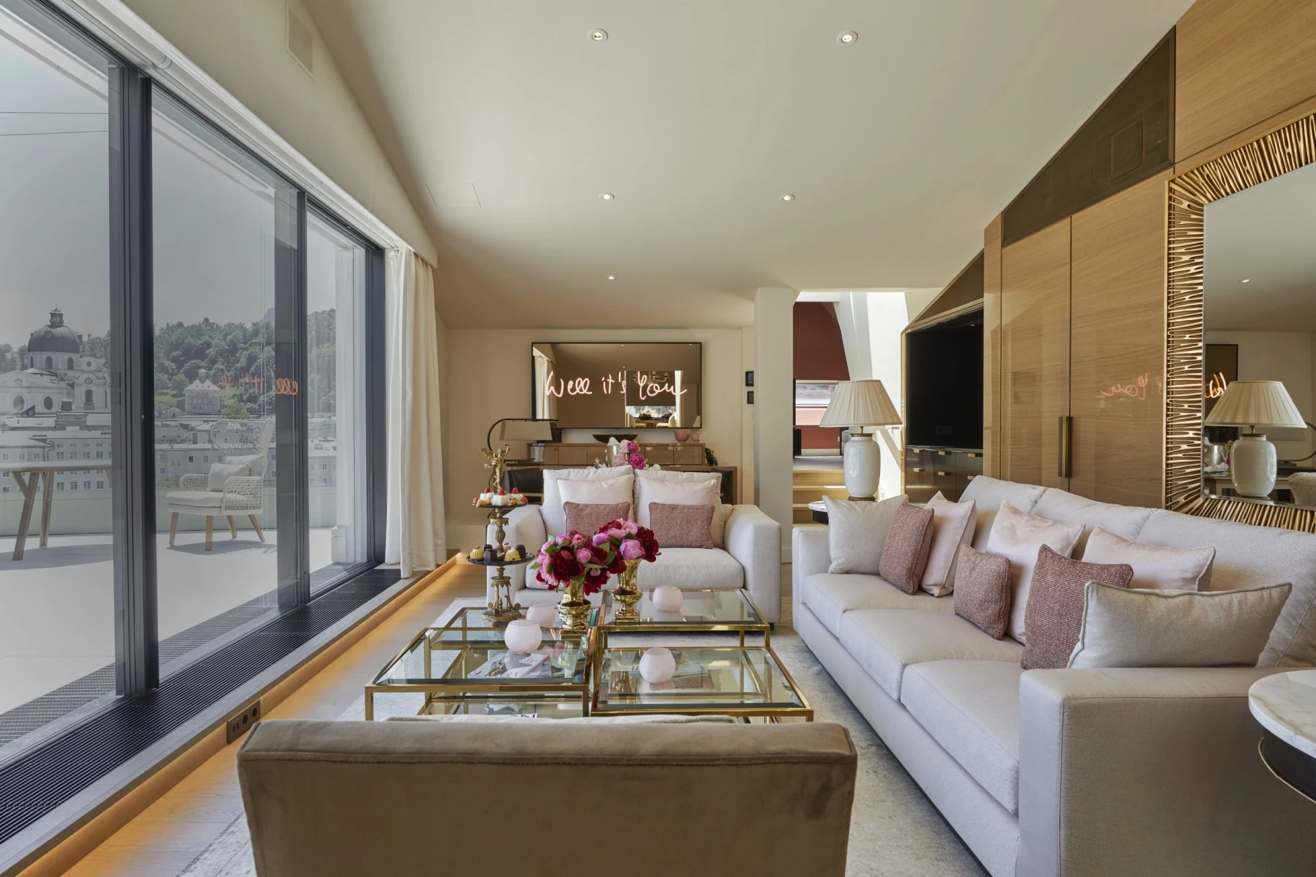 This is the elegant Sky Suite in Salzburg, a luxurious, light-filled living space with plush seating, gold-accented decor, fresh flowers, and sweeping panoramic views of the historic city through floor-to-ceiling windows.