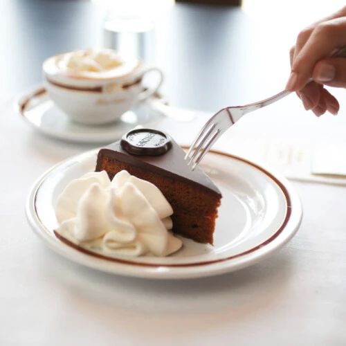 A hand reaches for a forkful of the iconic Original Sacher-Torte, elegantly served with whipped cream and accompanied by a creamy coffee on classic Hotel Sacher porcelain.