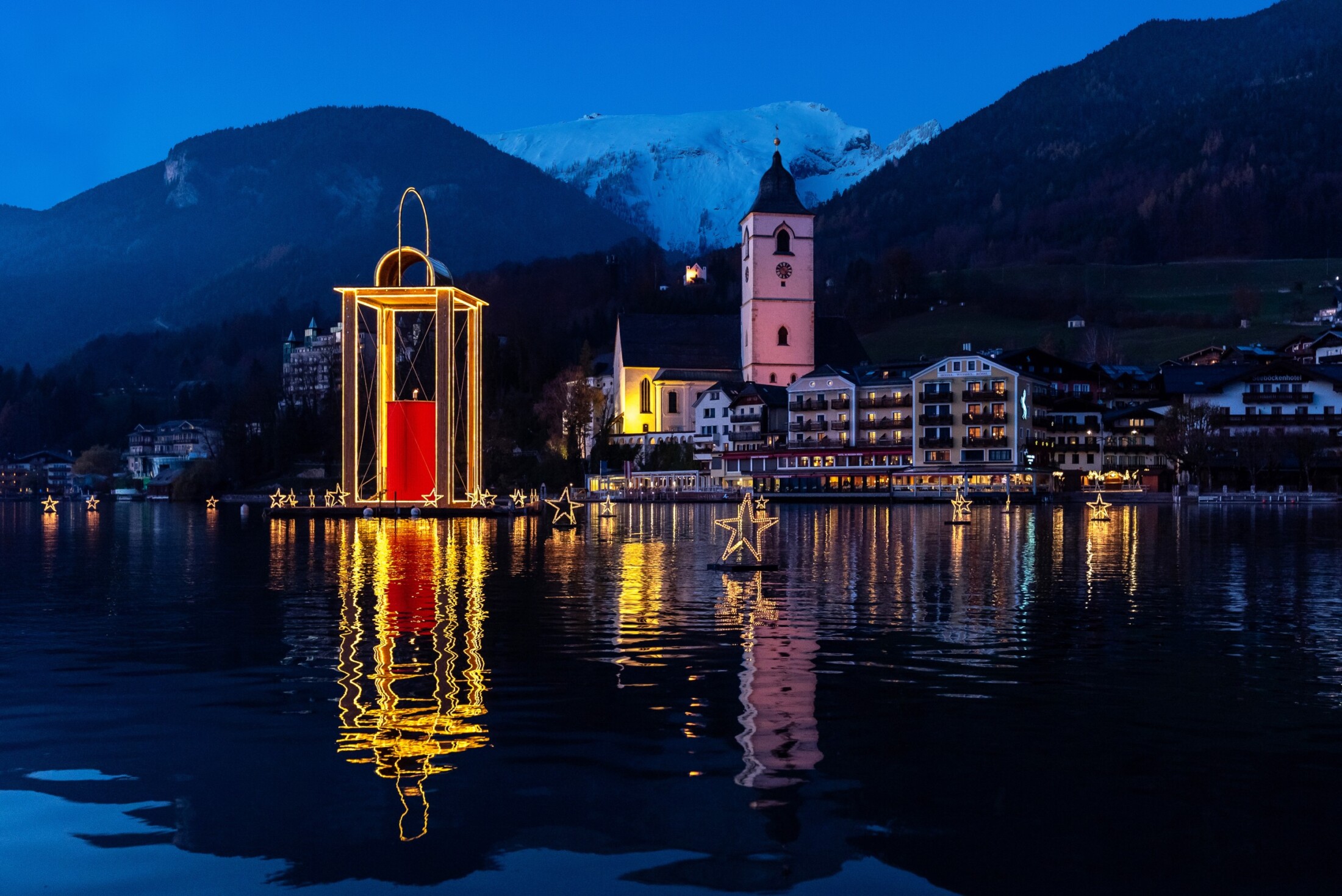 A magical evening scene in Salzburg features a glowing Christmas lantern and floating stars on the lake, with charming buildings and snow-capped mountains creating a festive alpine backdrop.