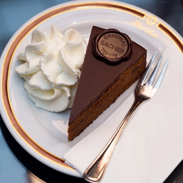 A decadent slice of the Original Sacher-Torte, topped with the signature chocolate seal, is served with a generous swirl of whipped cream on fine Hotel Sacher porcelain.