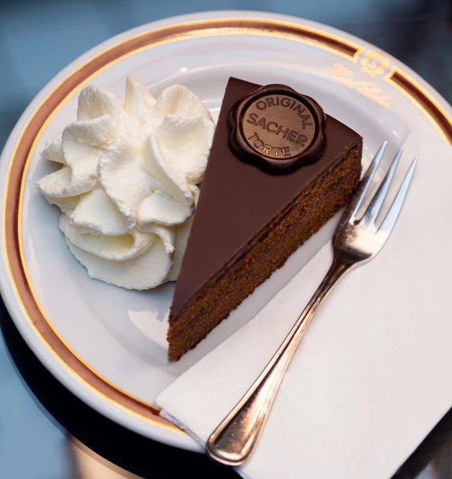 A decadent slice of the Original Sacher-Torte, topped with the signature chocolate seal, is served with a generous swirl of whipped cream on fine Hotel Sacher porcelain.