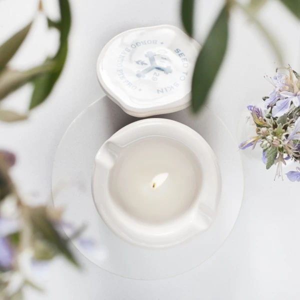The Massage Candle 2 A soothing white Seed to Skin candle burns gently in an elegant ceramic holder, surrounded by soft floral accents that enhance its calming ambiance.