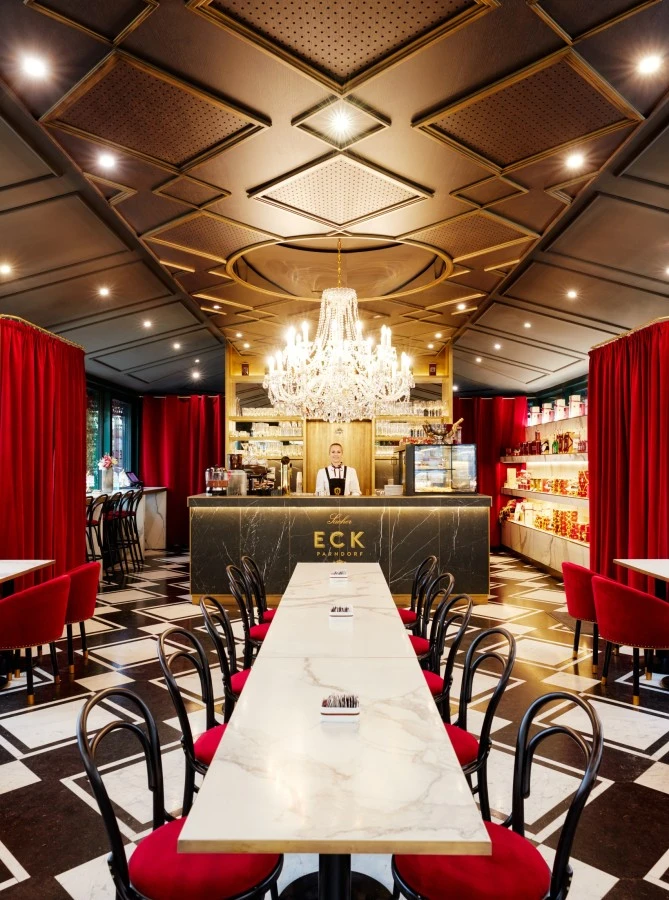 211008_JG-2262 Café Sacher Parndorf exudes elegance with its marble tables, red velvet chairs, dramatic chandeliers, and luxurious gold-accented bar under a richly designed ceiling.