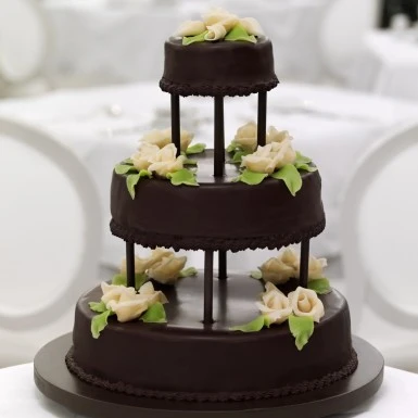 A three-tiered Original Sacher-Torte, coated in rich chocolate glaze and adorned with delicate white icing roses and green leaves, stands elegantly in a refined white setting.