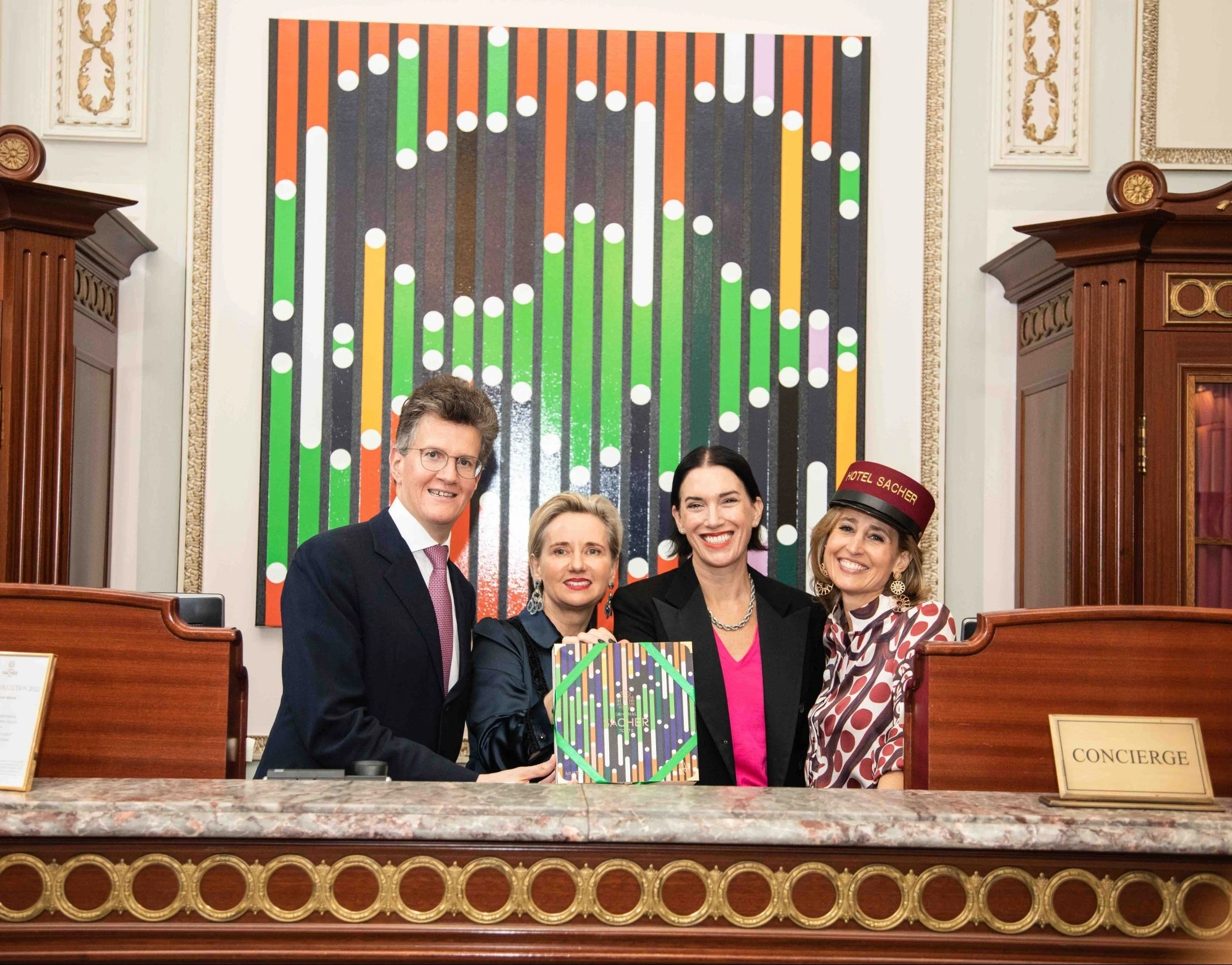 At the Hotel Sacher concierge desk, Sarah Morris, Alexandra Winkler, and guests proudly present the **Original Sacher-Torte Artist’s Collection 2022**, with the vibrant artwork featured prominently in the background.