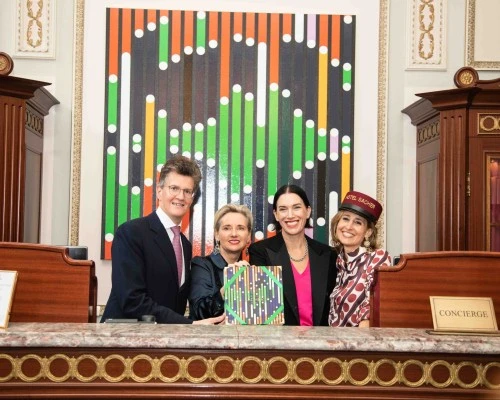 At the Hotel Sacher concierge desk, Sarah Morris, Alexandra Winkler, and guests proudly present the **Original Sacher-Torte Artist’s Collection 2022**, with the vibrant artwork featured prominently in the background.